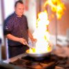 Uno chef in divisa cucina con una padella da cui si sprigiona una grande fiamma.
