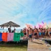 Persone su un molo festeggiano lanciando polveri colorate, sotto un cielo azzurro.