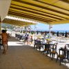 Area coperta con tavolini vicino alla spiaggia, persone passeggiano, vista del mare e ombrelloni.