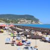 Spiaggia sabbiosa con ombrelloni scuri, mare blu e colline sullo sfondo.