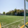 Un campo di calcio con erba sintetica e linee gialle, recintato, con alberi intorno e cielo sereno.